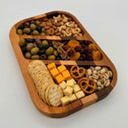 Wooden snack platter with a variety of snacks including olives, nuts, dried fruits, crackers, and cheese on a white background.